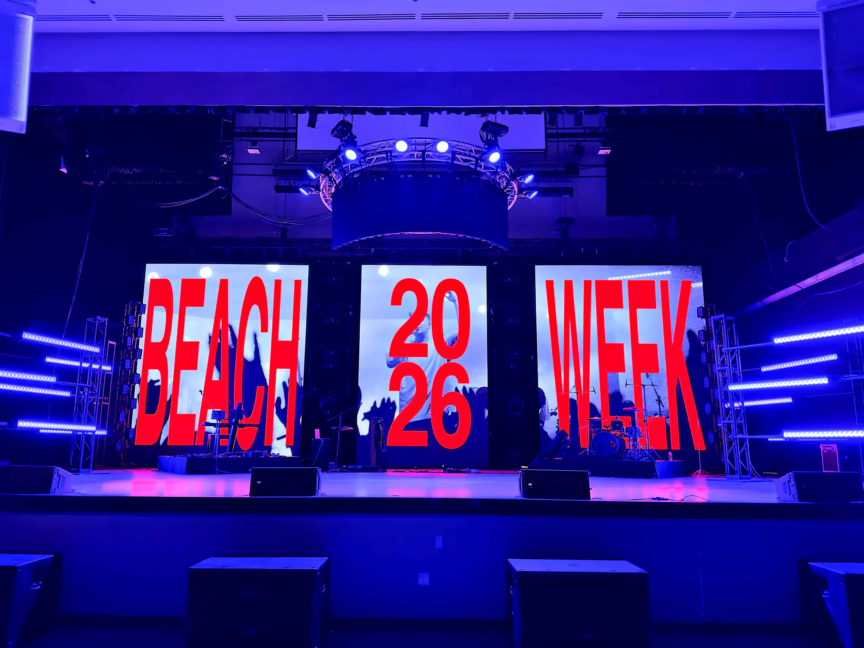 Beach Week 2026 stage at Arnold High School theater: three-panel LED wall, curved truss halo, and color-bar lighting towers
