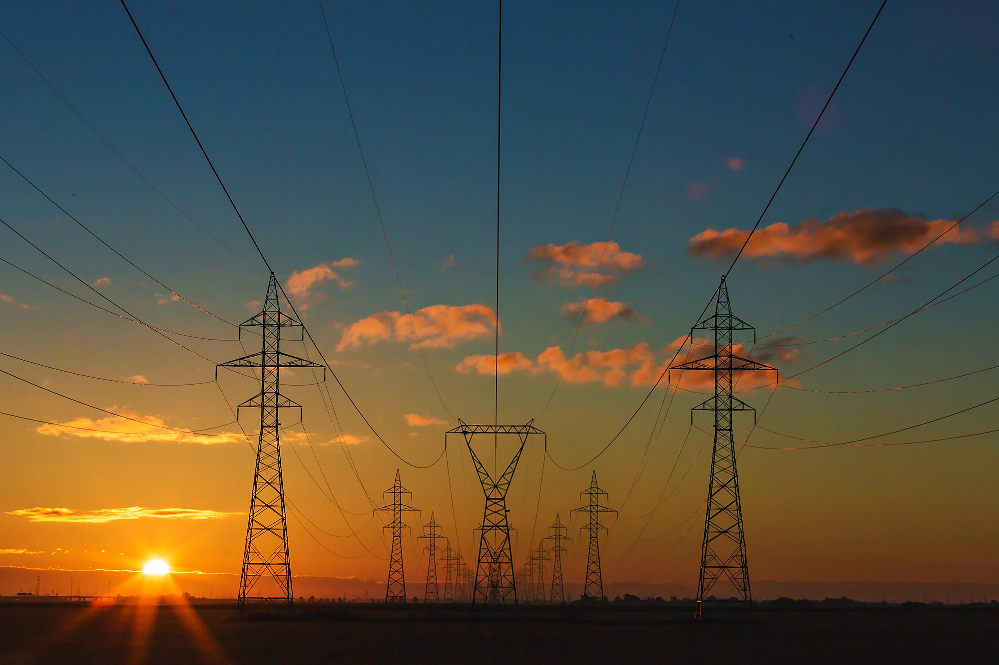 High-voltage transmission towers at dusk, the distributed infrastructure an energy broadcast has to reach