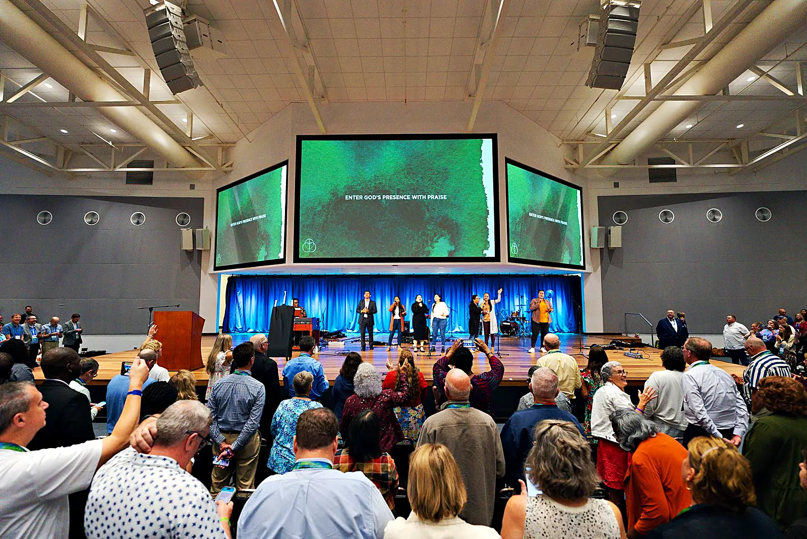 The full conference hall mid-worship: three screens, stage band, standing congregation