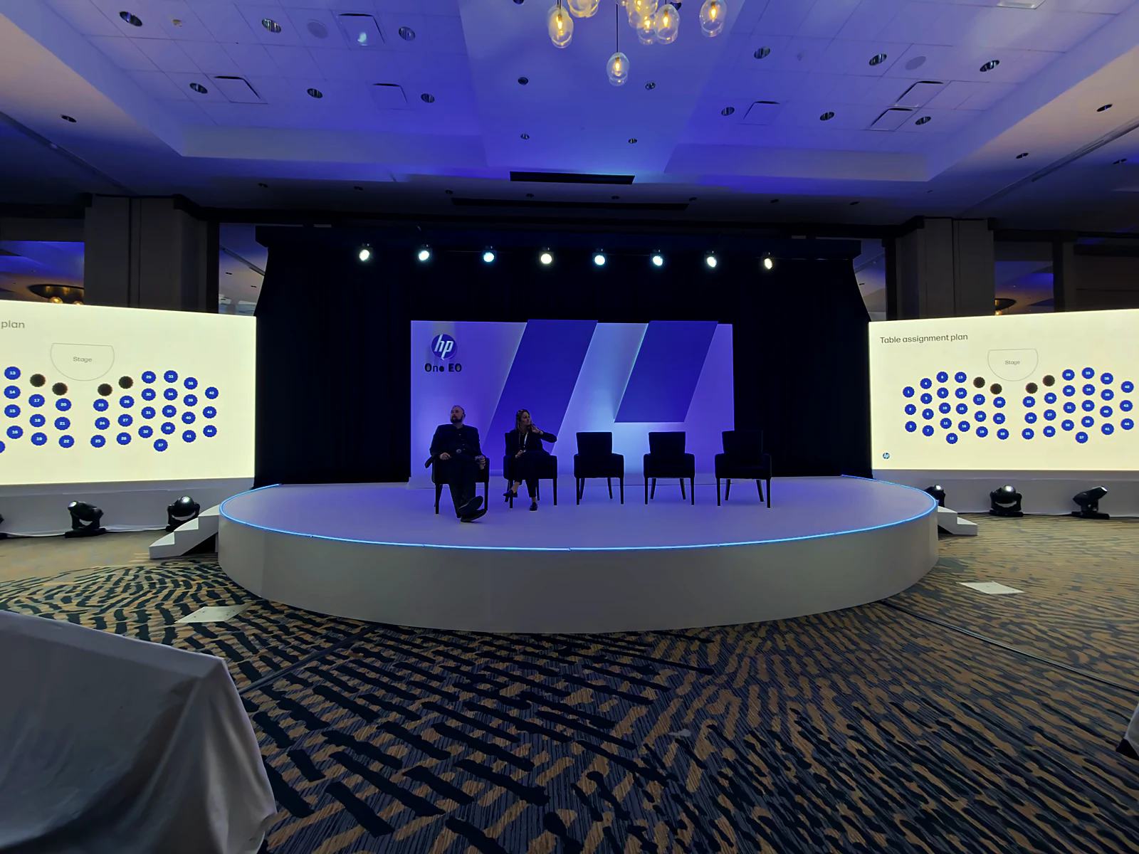 Fireside panel seated on the half-circle stage under blue light, table-assignment maps on the side screens