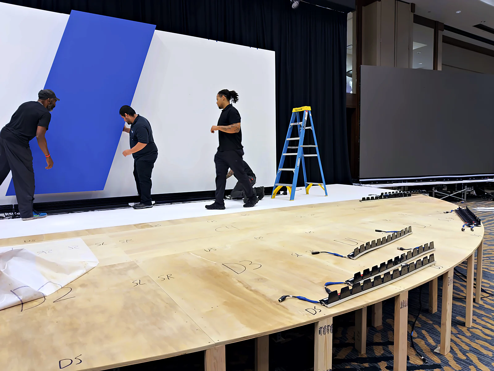 Crew installing the scenic wall behind the raw curved half-circle stage deck with position markers
