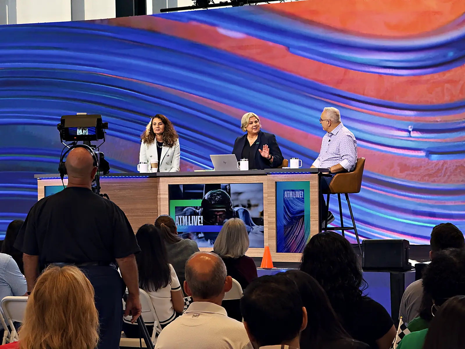 Three panelists live at the branded anchor desk, broadcast camera in the foreground